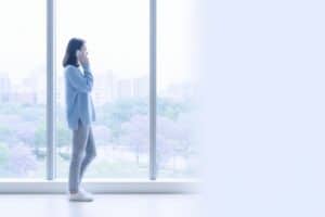 Una mujer joven de perfil habla por teléfono móvil de pie junto a un gran ventanal con vistas a la ciudad.